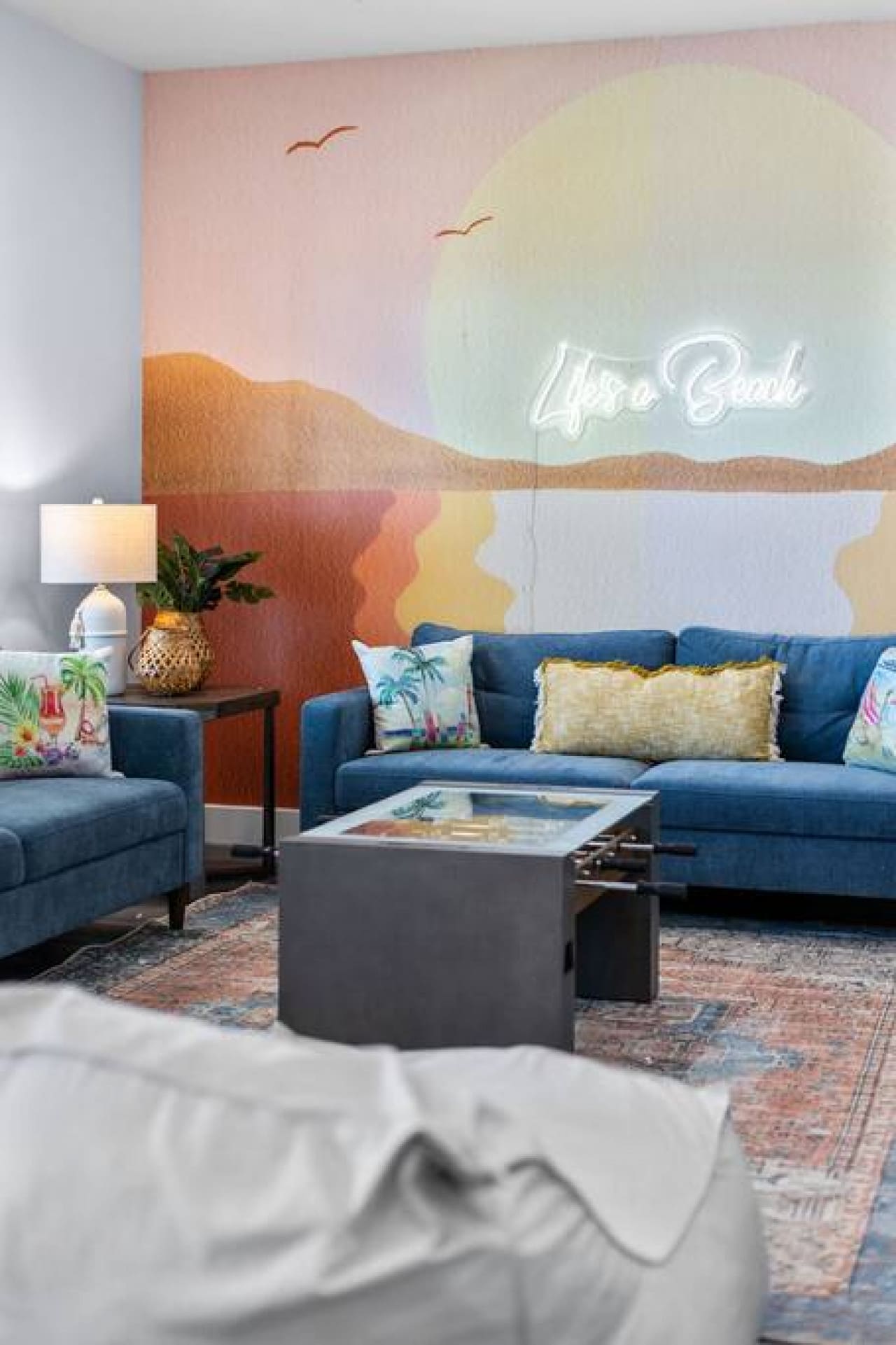 A serene beach-inspired Airbnb interior design with 'Lyra Beach' neon sign and sunset wall mural.